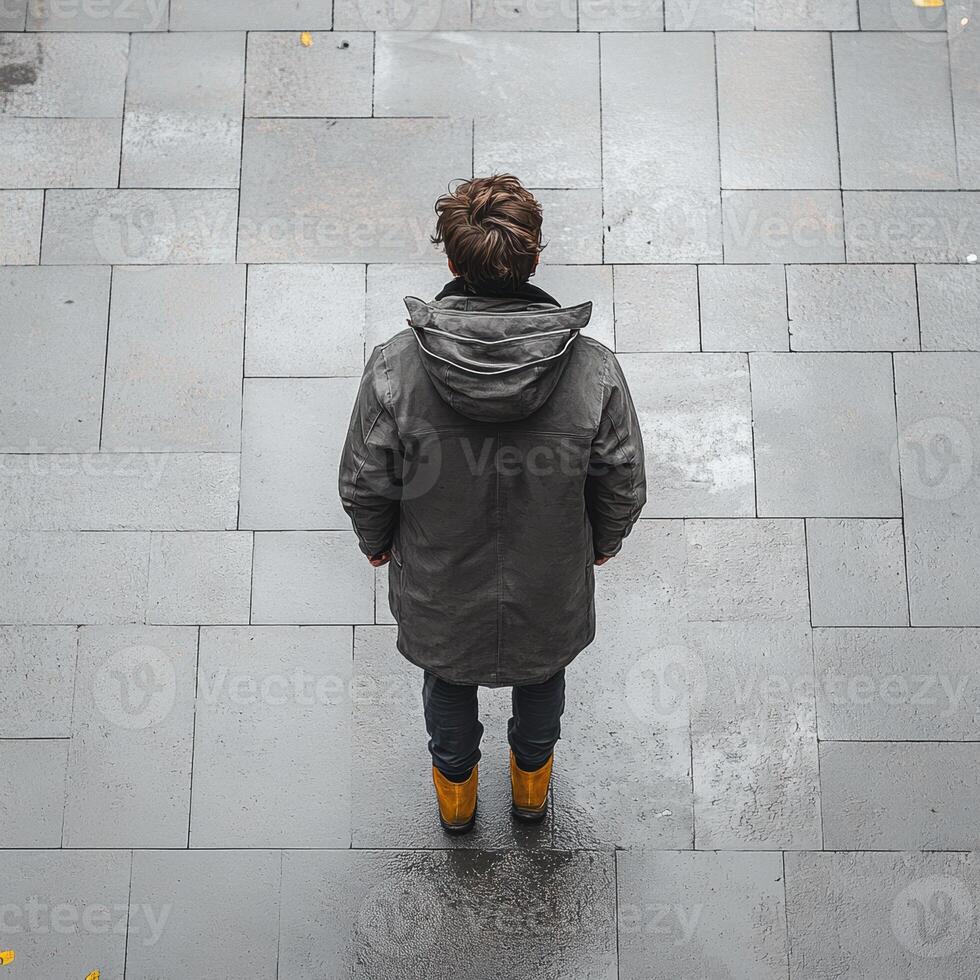 man in dark coat stands on wet pavement, facing away, showcasing stylish look. scene captures moment of contemplation and solitude photo