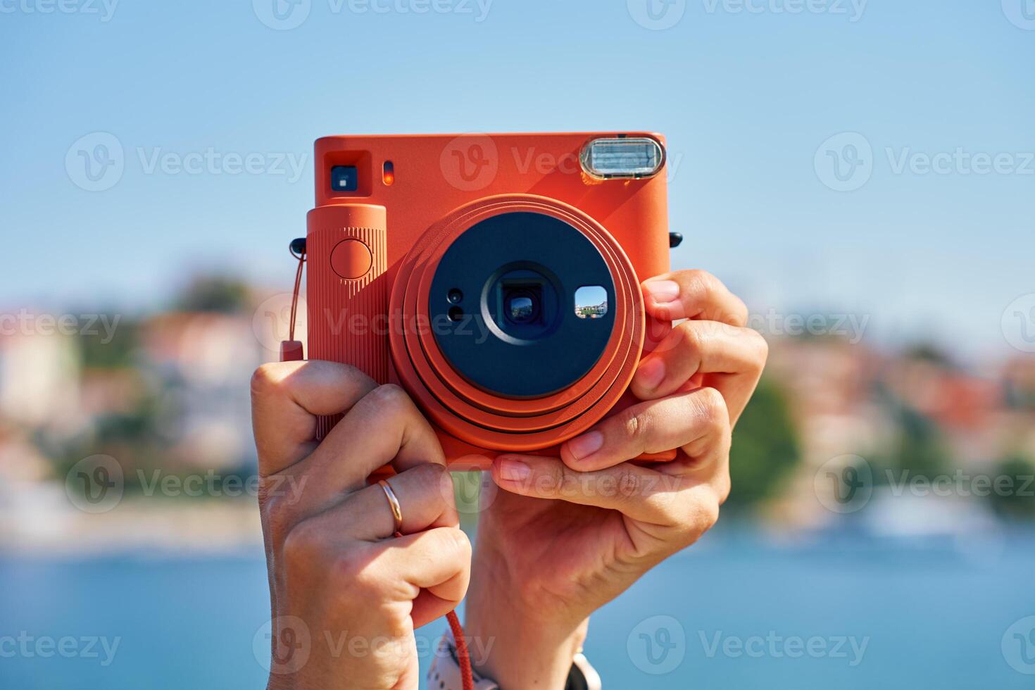 Close-up of tourist hand holding an orange instant camera in retro style with coastal town and blue sea in background. Summer travel concept. Equipment for capturing moments photo