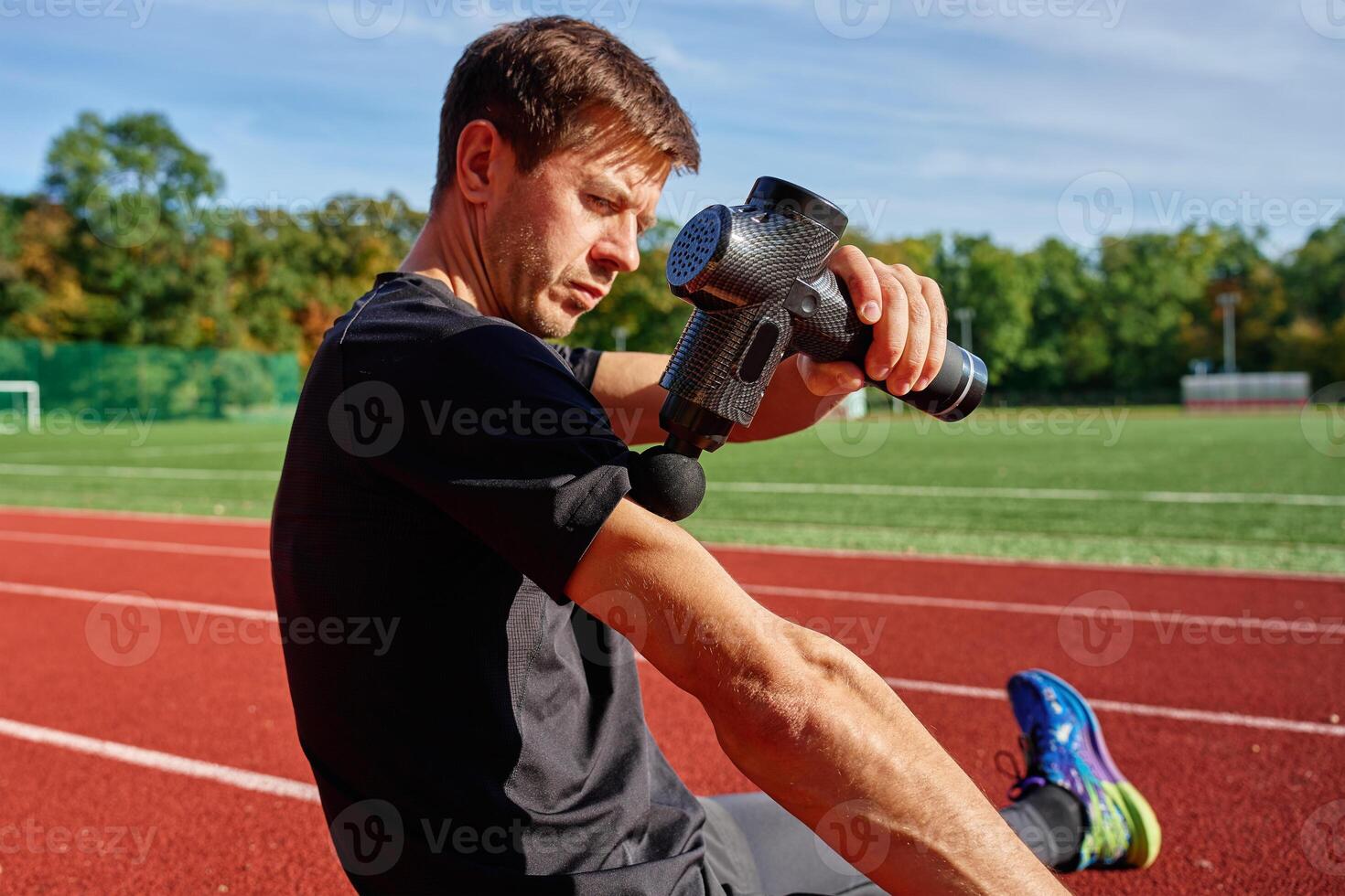 Close-up of male athlete using handheld massage gun on arm muscles for recovery after training session on stadium track. Concept of muscle relaxation and sports recovery photo