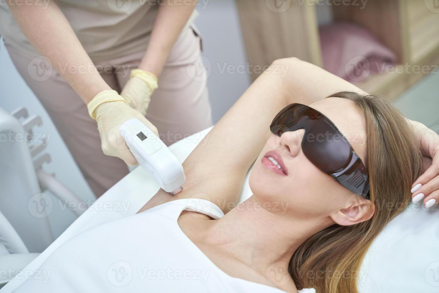 Woman receiving laser hair removal treatment on armpit in a clinic setting with protective eyewear. photo
