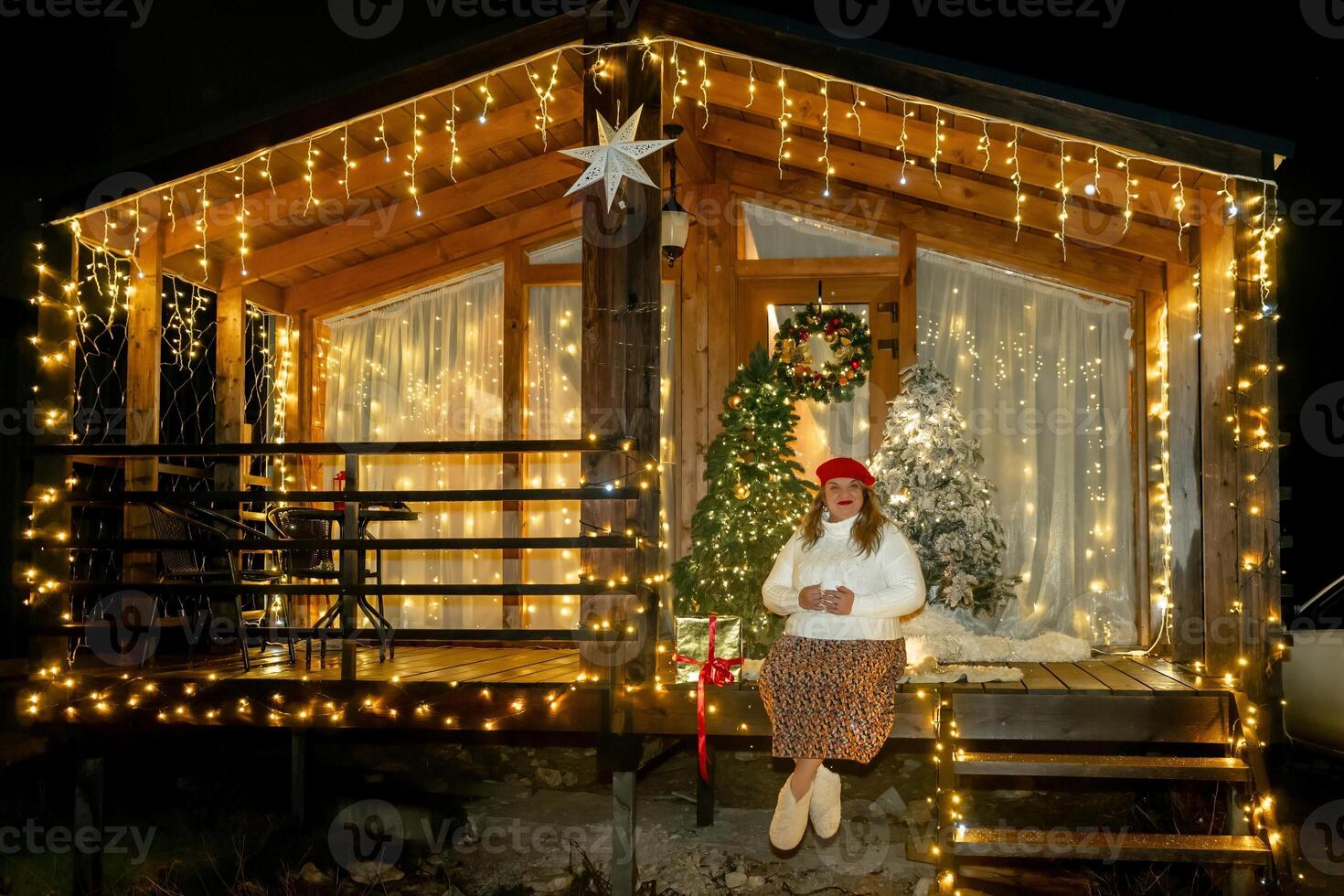 Woman Christmas Lights Cabin - A woman wearing a Christmas hat stands outside a cabin decorated with Christmas lights at night. photo