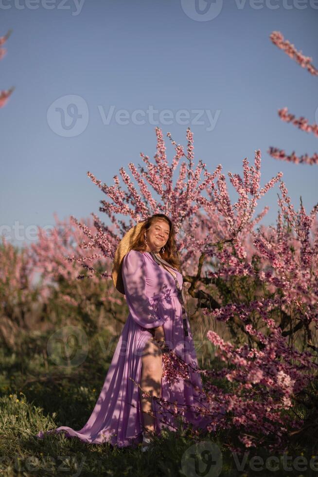 Woman blooming peach orchard. Against the backdrop of a picturesque peach orchard, a woman in a long pink dress and hat enjoys a peaceful walk in the park, surrounded by the beauty of nature. photo