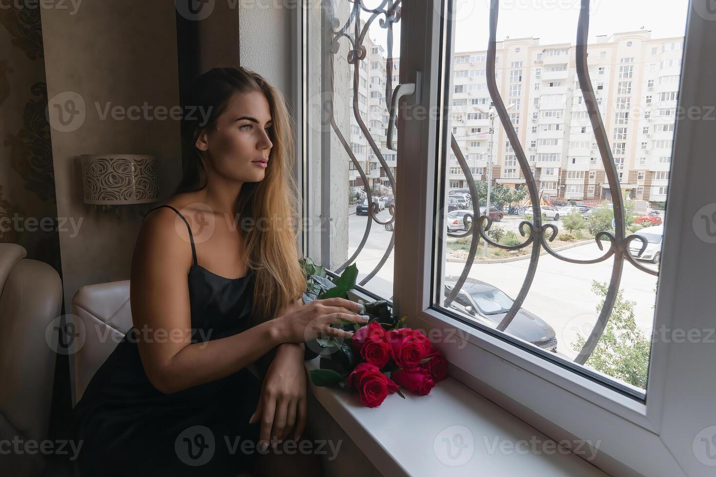A woman in a black dress is sitting in front of a window with a bouquet of red roses. photo
