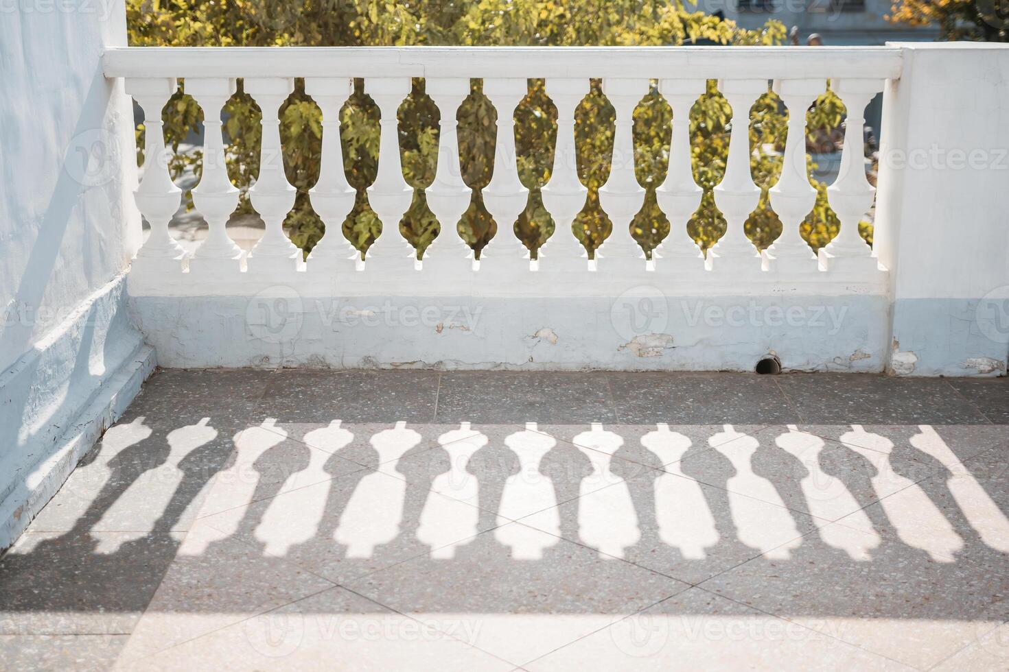A balcony with a railing and a tree in the background. The railing is white and has a shadow on it. photo