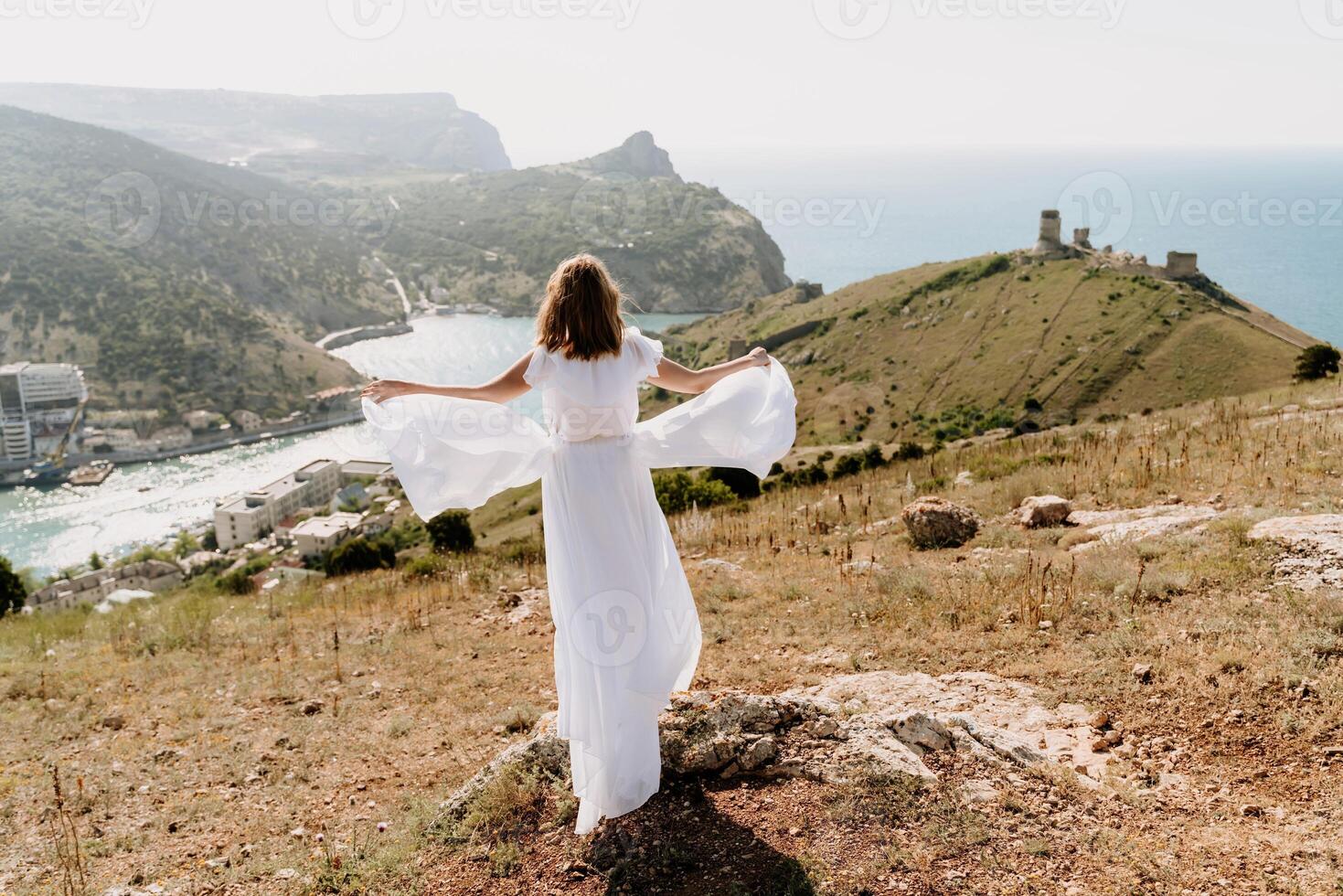 Happy woman in a white dress and hat stands on a rocky cliff above the sea, with the beautiful silhouette of hills in thick fog in the background. photo