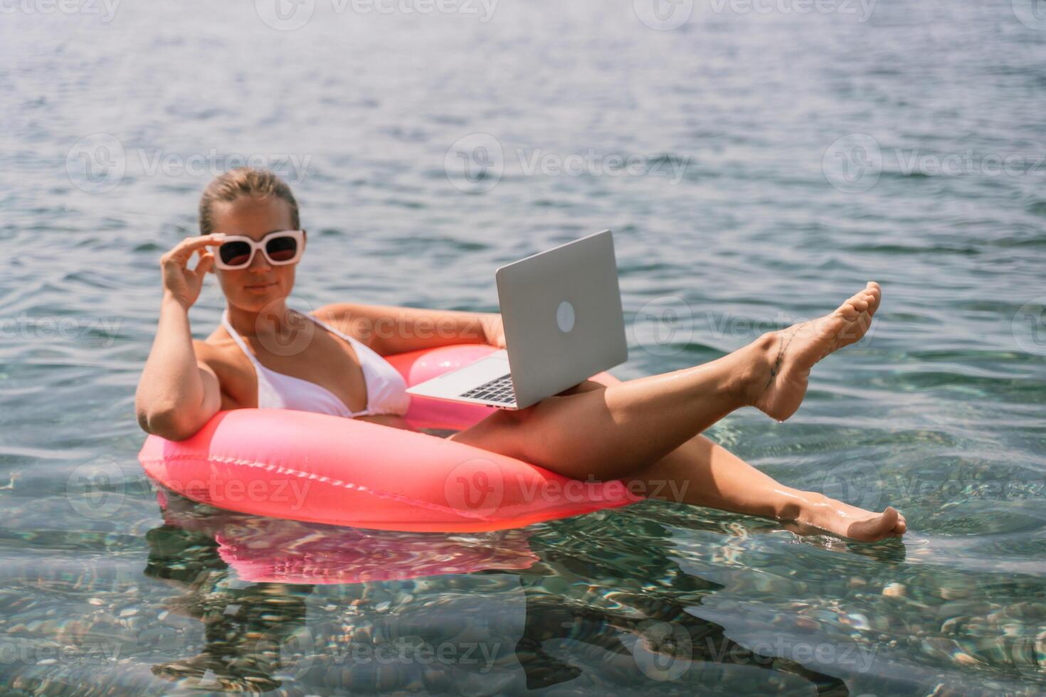 A woman is floating in a pink inflatable raft with a laptop in her hand. She is smiling and she is enjoying her time in the water. photo