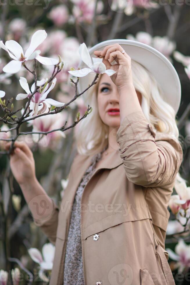 Magnolia flowers woman. A blonde woman wearing a white hat stands in front of a tree with pink flowers. She has a smile on her face and she is enjoying the beautiful scenery. photo