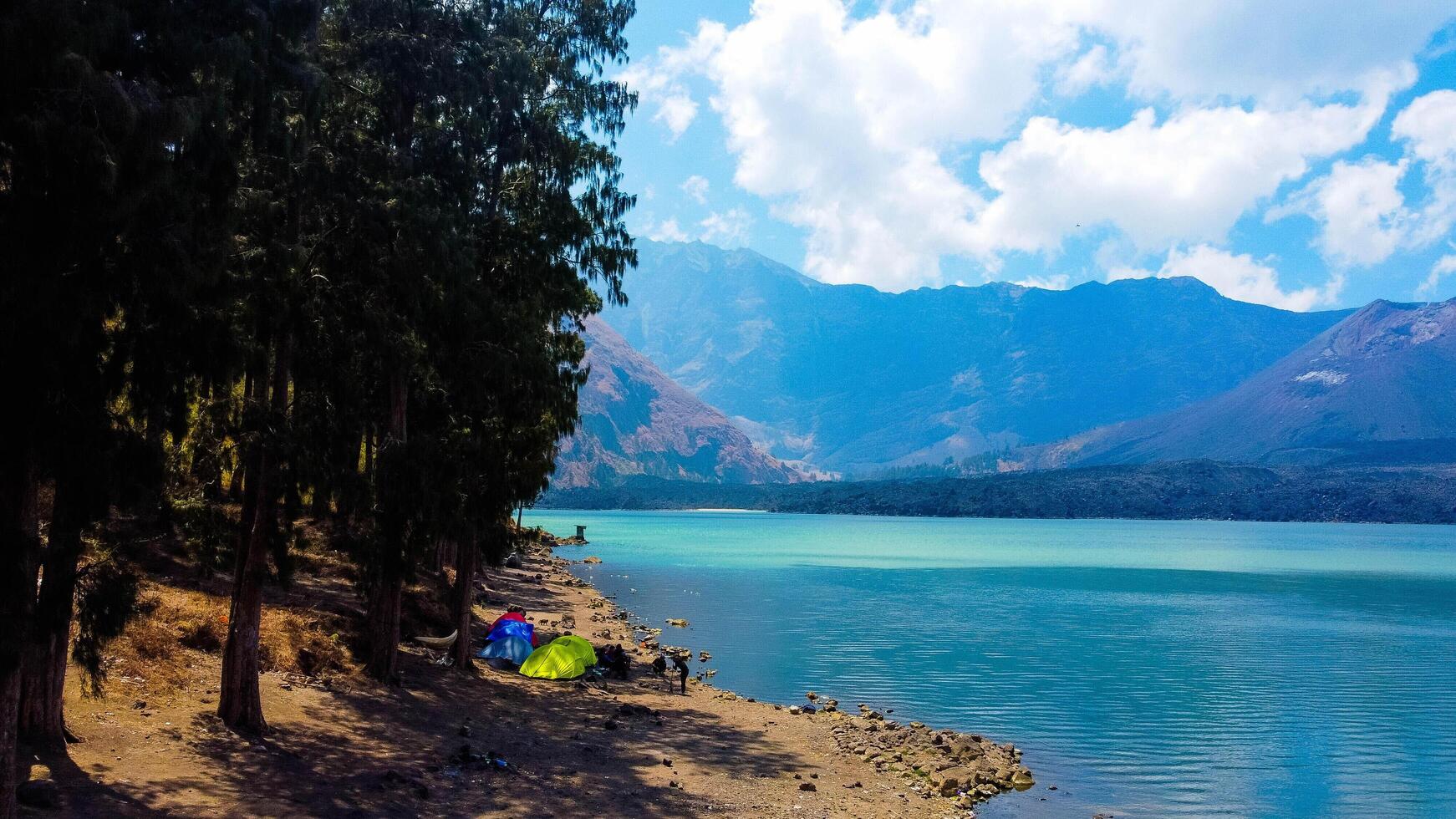 Aerial from the summit of Rinjani shows the stunning Segara Anak Lake below, Surrounded by vibrant blue skies and clouds, the lake's serene beauty stands out against the dramatic landscape. photo