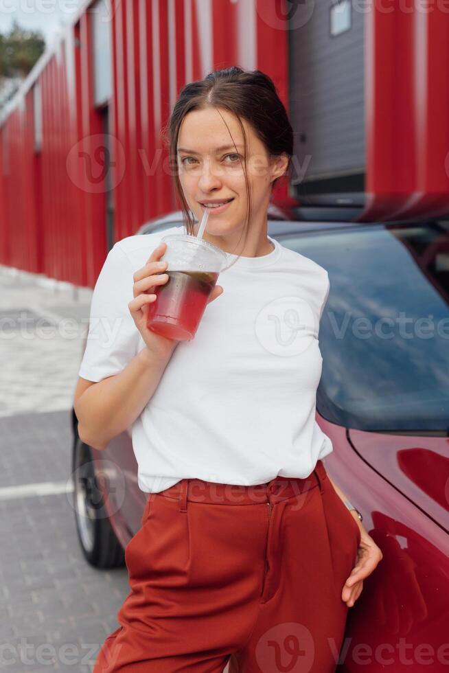 Attractive woman in stylish outfit drinks fresh lemonade near red car on a vibrant red urban backdrop photo