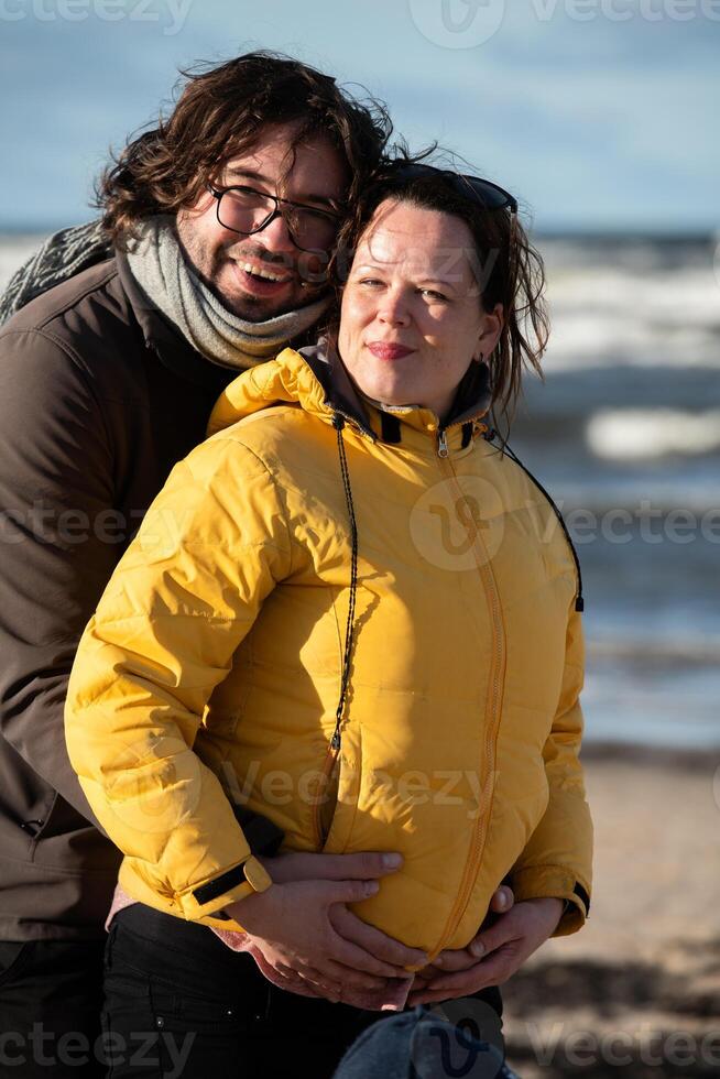 A couple stands embraced on a sandy beach on a chilly autumn day, the serene ocean stretching behind them as they share a quiet moment of love and warmth amidst the cool breeze and peaceful photo