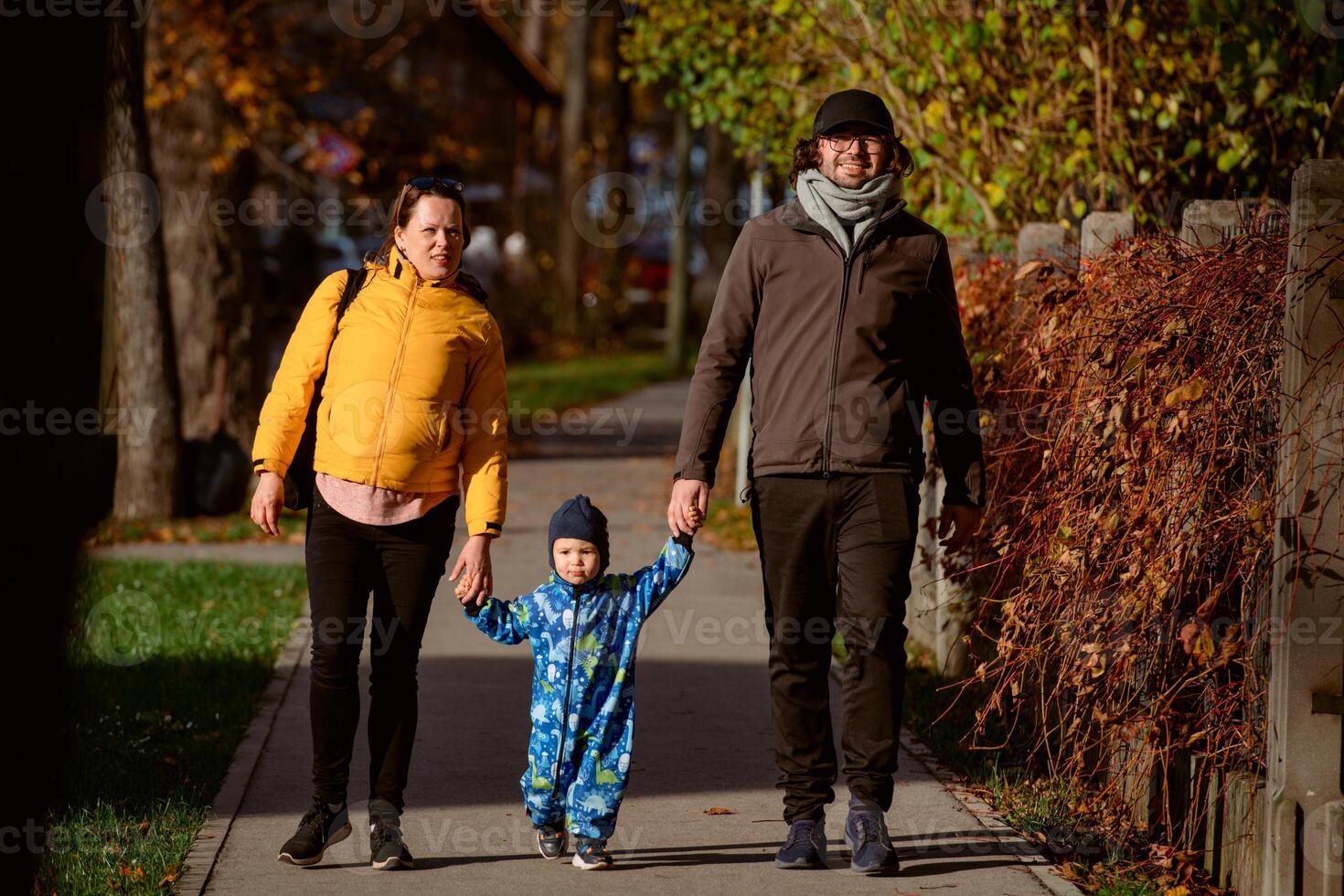 A loving couple strolls through a sunlit park with their young son, surrounded by the vibrant colors of autumn, enjoying a joyful and peaceful family moment together. photo