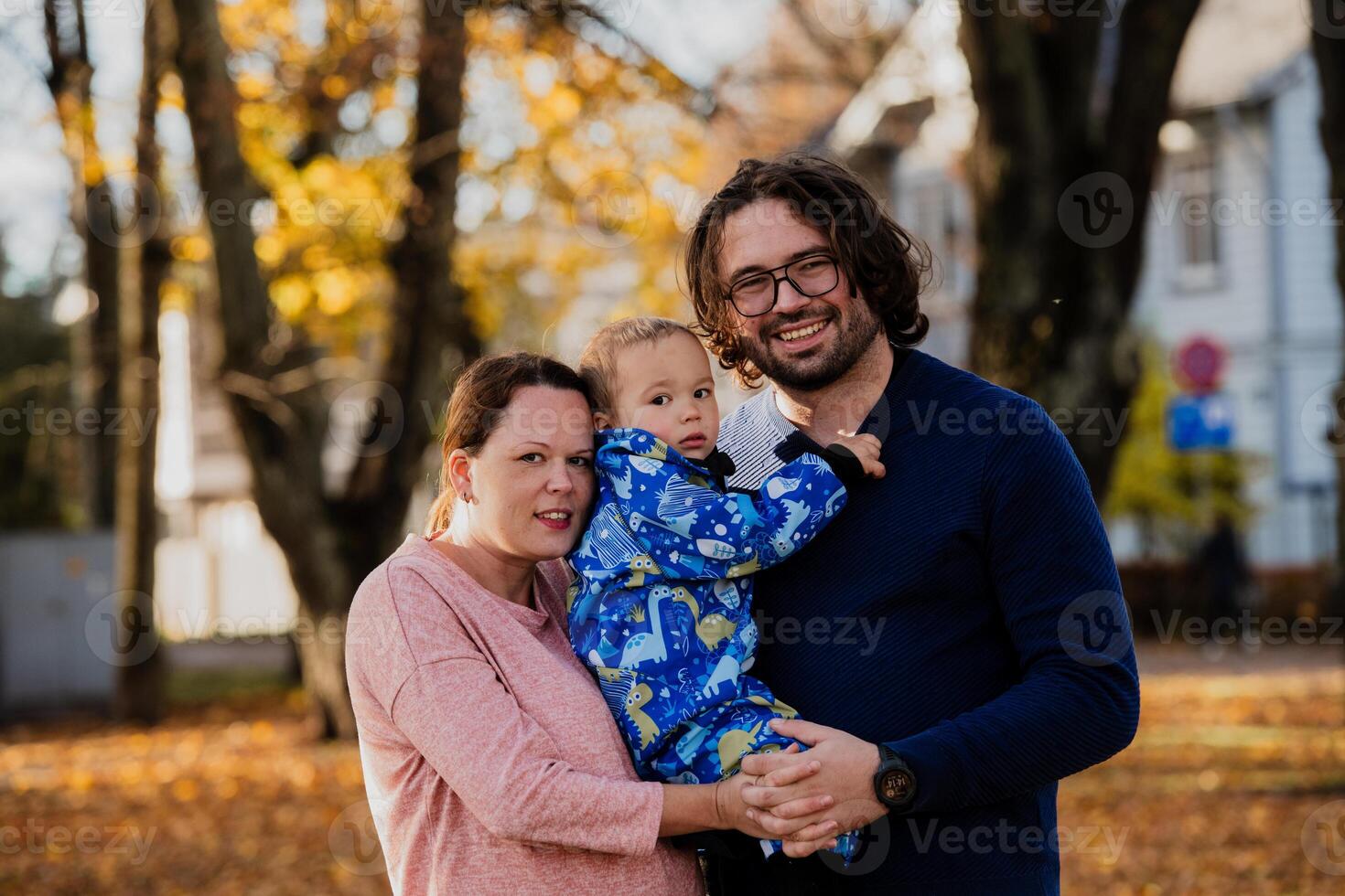 On a sunny autumn day in the park, a mother and father lovingly embrace their young son, sharing moments of laughter and joy as they play together photo