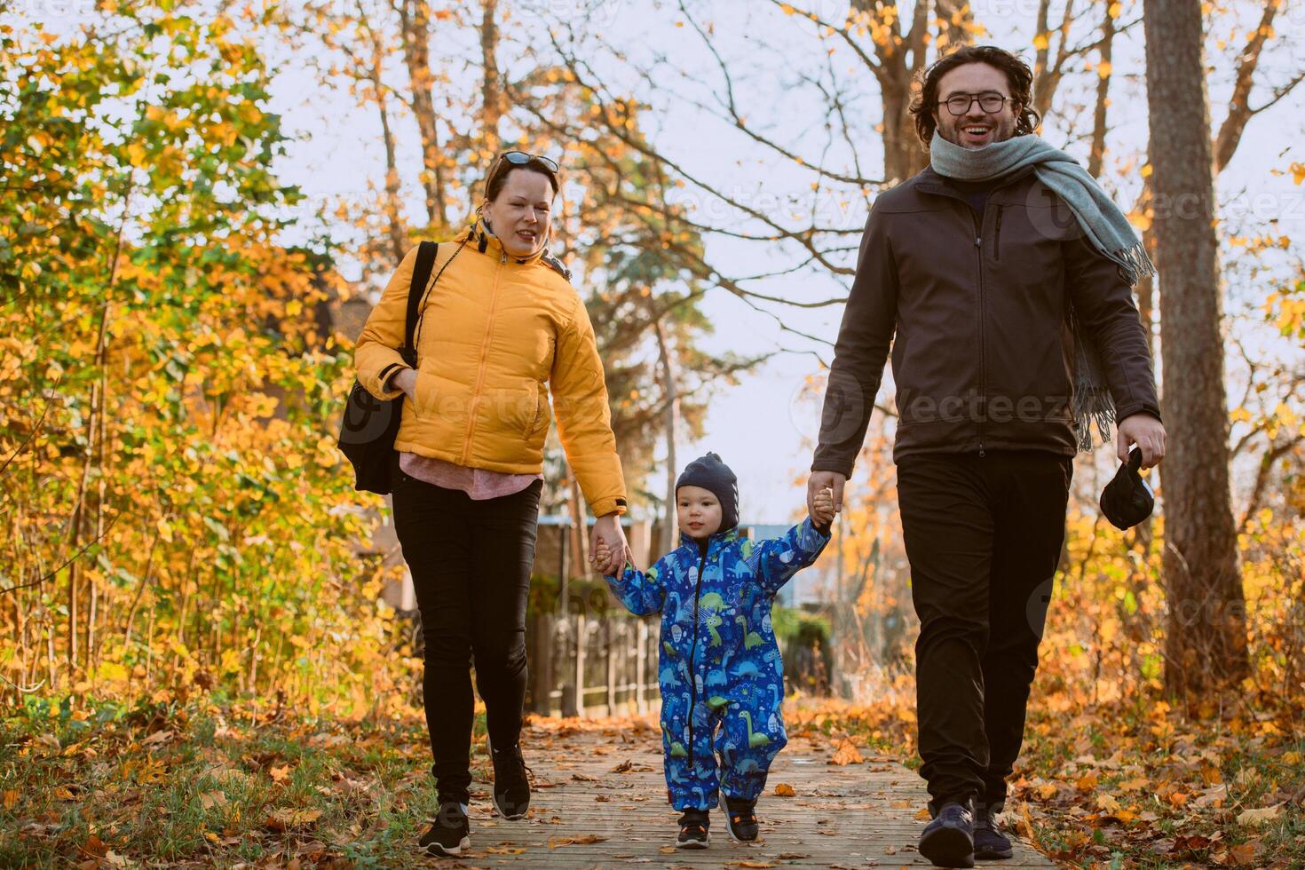 A loving couple strolls through a sunlit park with their young son, surrounded by the vibrant colors of autumn, enjoying a joyful and peaceful family moment together. photo