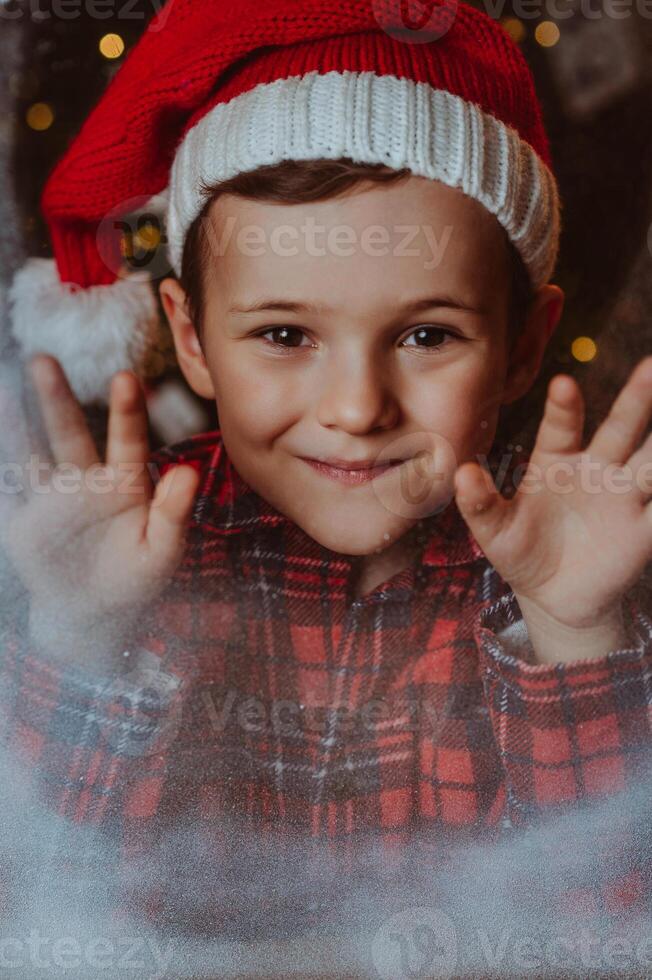 the joyful face of a boy in a Santa hat at a snow-covered window. Festive mood. photo