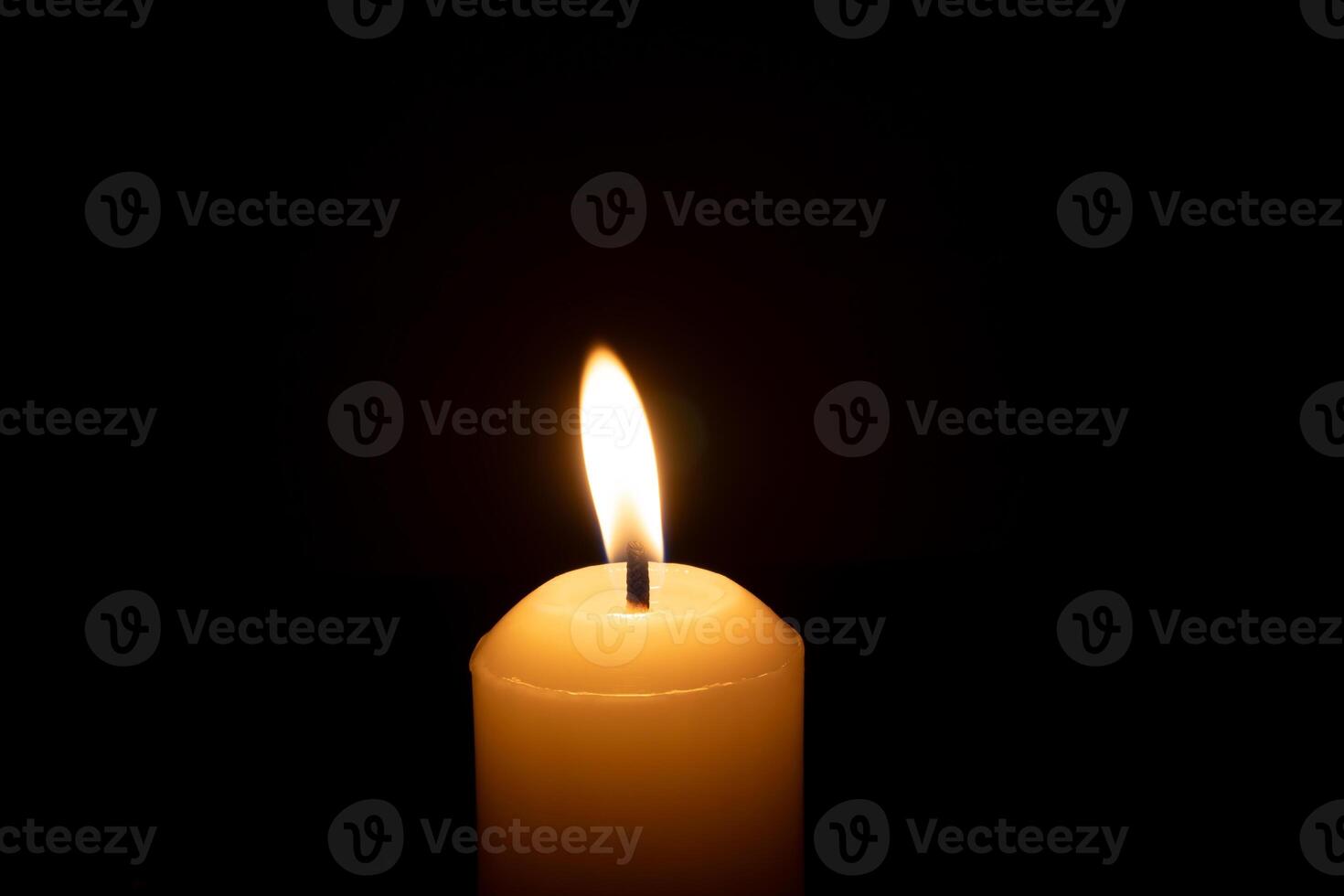 A single burning candle flame or light glowing on a big white candle isolated on black or dark background on table in church for Christmas, funeral or memorial service photo