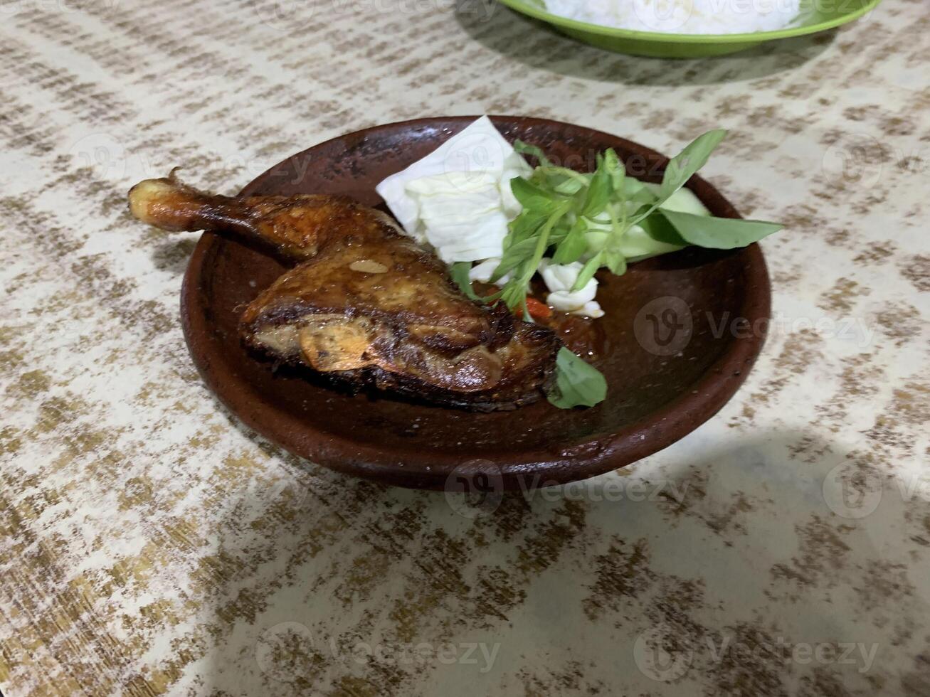Ayam bakar Penyet is a fried chicken served with hot chili in a plate with fresh vegetables, slices of cucumber and basil leaves photo