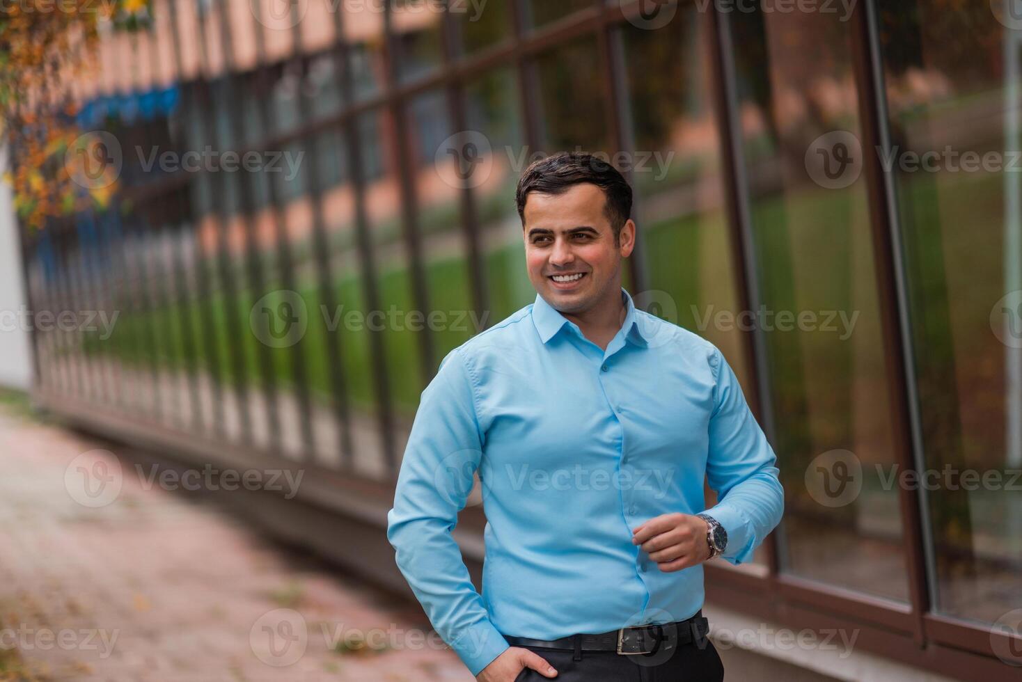 Portrait of a happy young businessman standing near the modern office building. A handsome confident young man standing looking at camera and smiling in a blue shirt outside. photo