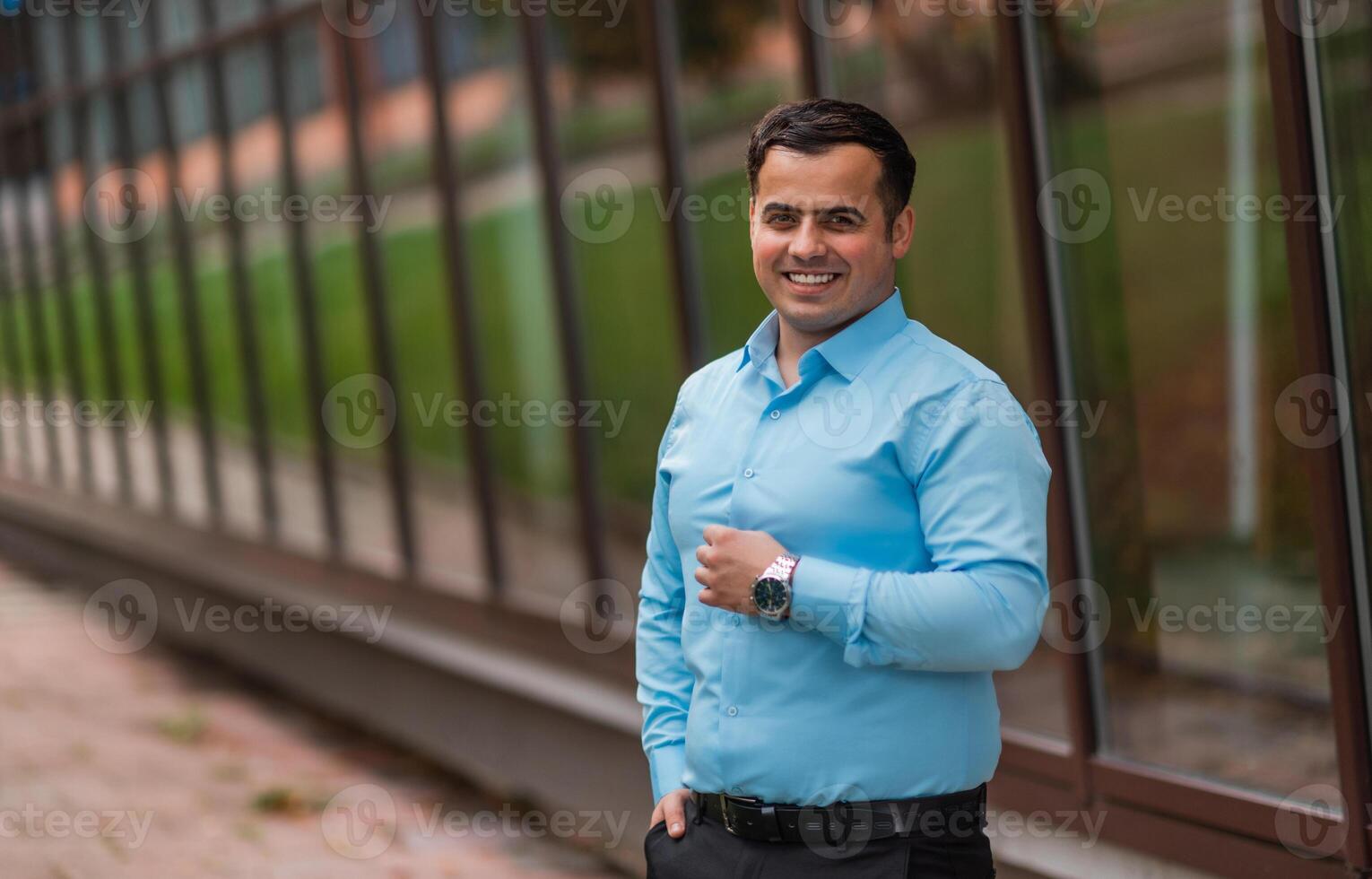 Portrait of a happy young businessman standing near the modern office building. A handsome confident young man standing looking at camera and smiling in a blue shirt outside. photo