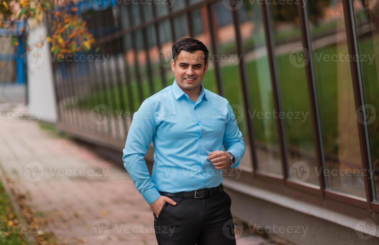Portrait of a happy young businessman standing near the modern office building. A handsome confident young man standing and smiling in a blue shirt outside. photo