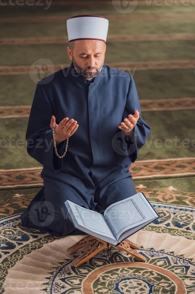 A Muslim prayer man praying with hands up while reading a Quran inside the mosque. photo