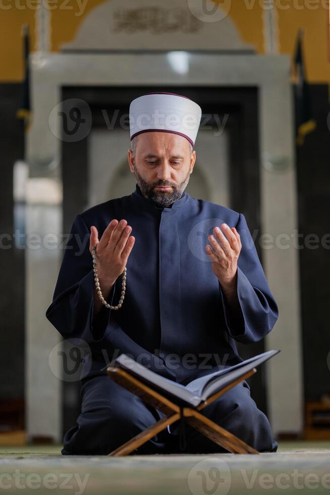 A Muslim prayer man praying with hands up while reading a Quran inside the mosque. photo