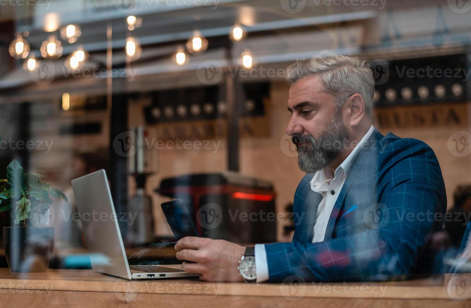 A handsome mature business man using his phone while working on a laptop in the office near the window. photo