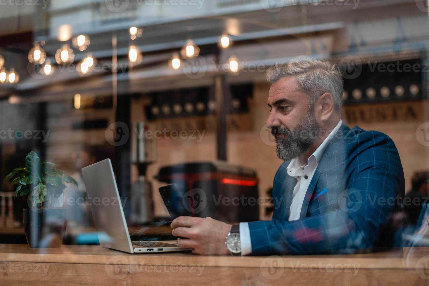 A handsome mature business man using his phone while working on a laptop in the office near the window. photo