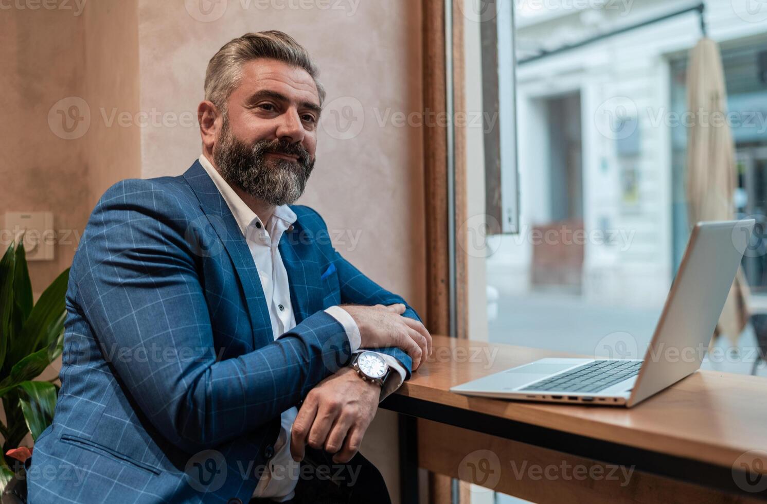 Portrait of a happy mature business man sitting at his desk in the office or restaurant cafe near the window while working on a laptop. photo
