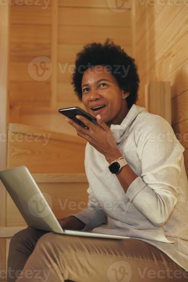 African Woman Sending Audio Message On A Smartphone While Working On Laptop photo