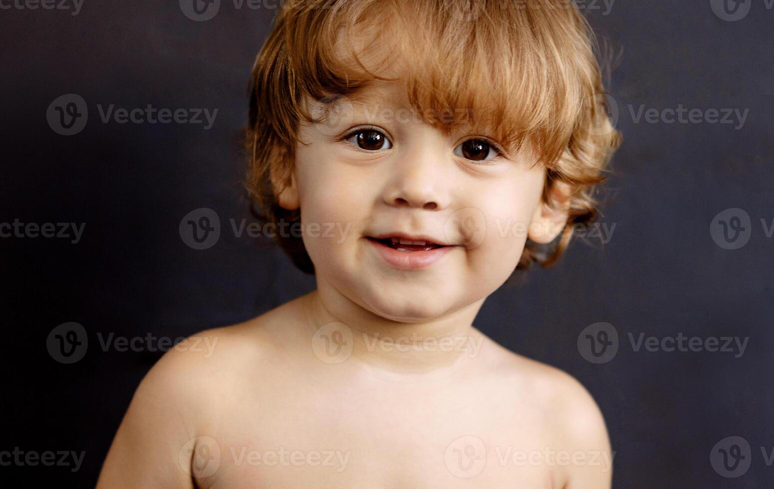 A close-up portrait of a smiling cute boy. A blond sweet little baby is looking at the camera. An adorable laughing kid in the studio. Black background. photo