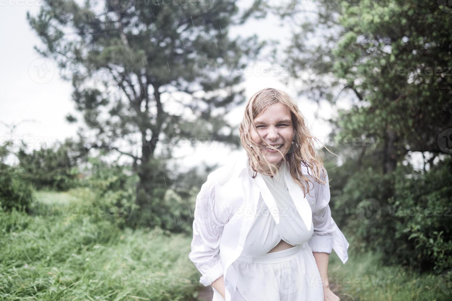 A close-up portrait of a laughing young woman in a white dress in the rain. A charming smiling girl enjoys the summer precipitation. A wet girl is having fun in the rain in the park. photo