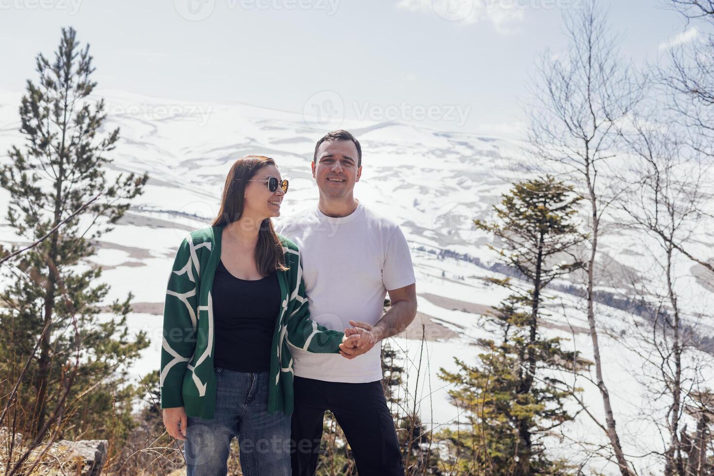 Young married couple in casual clothes stands and looks at snowy landscape. Smiling man and woman hug and enjoy views of mountains. photo