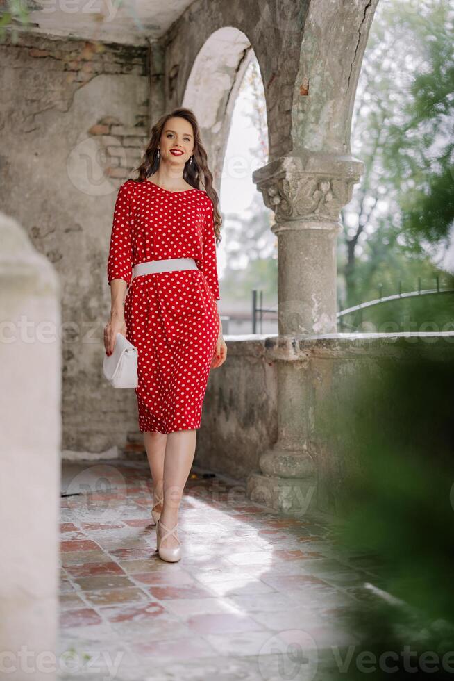 Elegant woman walking in red polka dot dress at ancient building archway photo