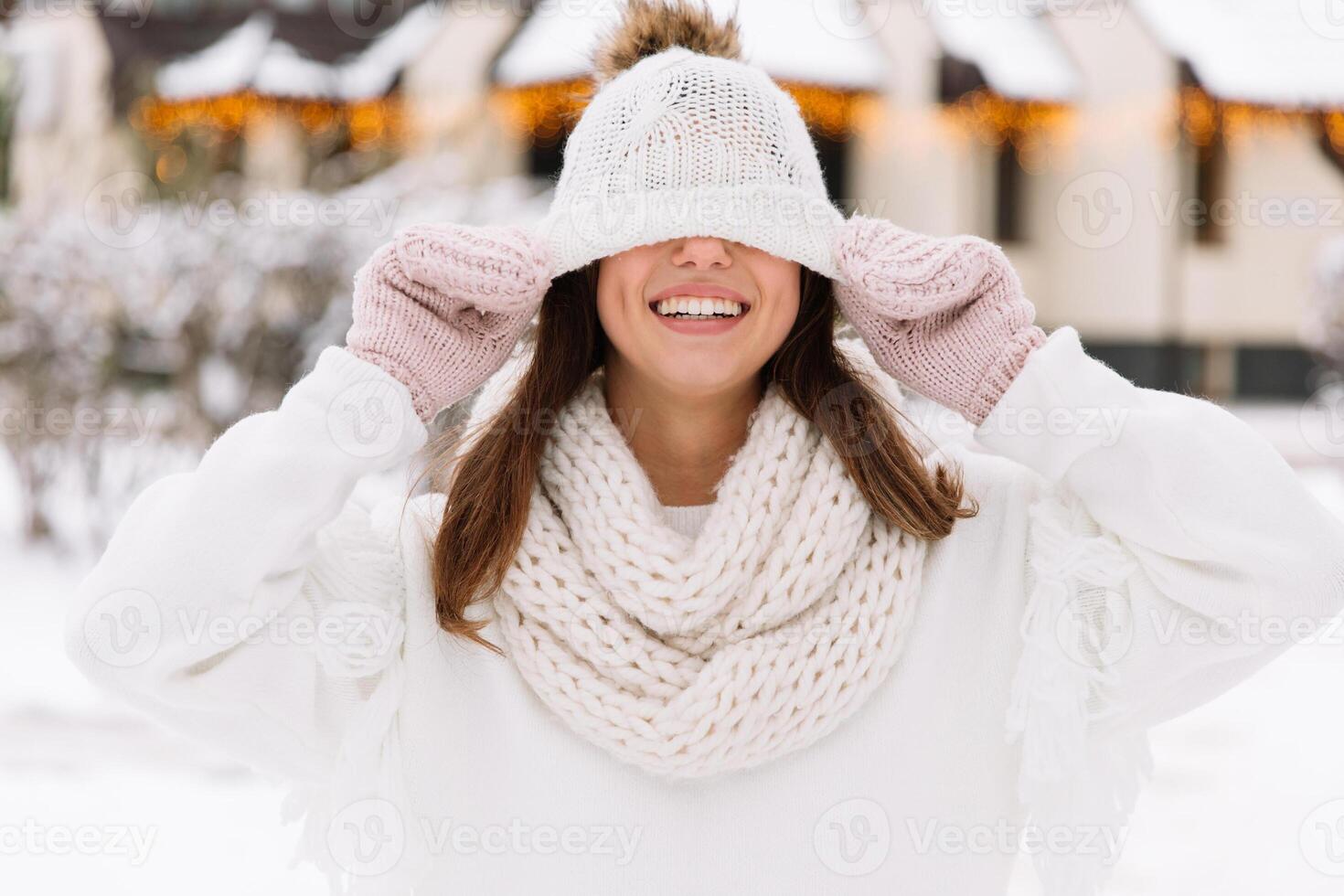 Outdoor close up portrait of young beautiful happy smiling girl wearing white knitted, scarf and gloves. The model jokes and closes the eyes with hat in park with Christmas lights. photo