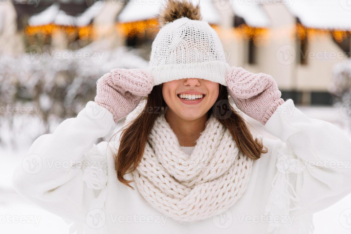 Outdoor close up portrait of young beautiful happy smiling girl wearing white knitted, scarf and gloves. The model jokes and closes the eyes with hat in park with Christmas lights. photo