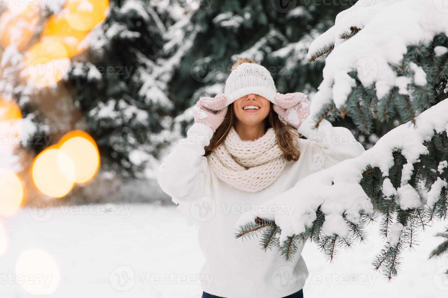 Outdoor close up portrait of young beautiful happy smiling girl wearing white knitted, scarf and gloves. The model jokes and closes the eyes with hat in park with Christmas lights. photo