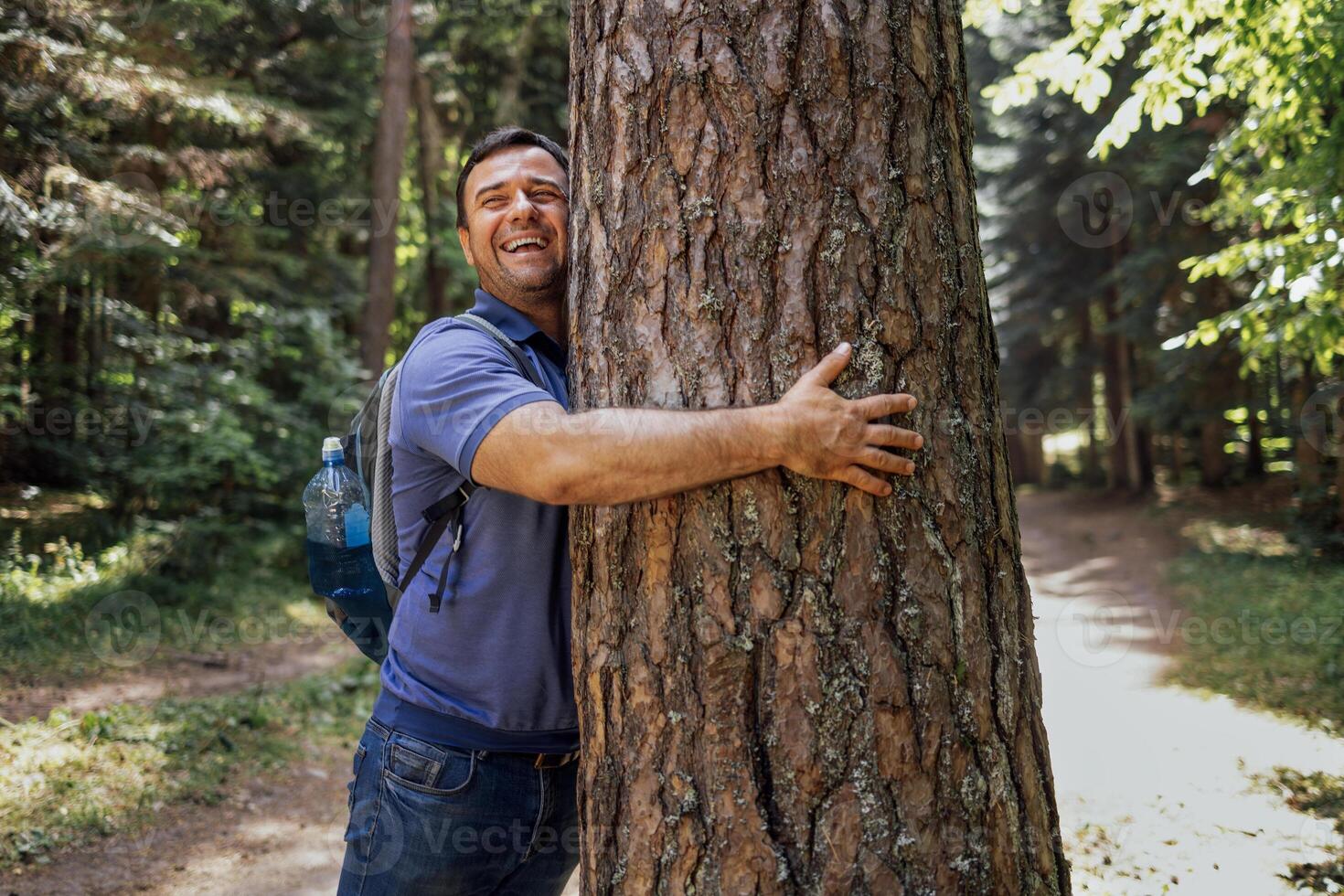 A young man with a backpack in the woods. An attractive tourist touches a large tree in the forest photo