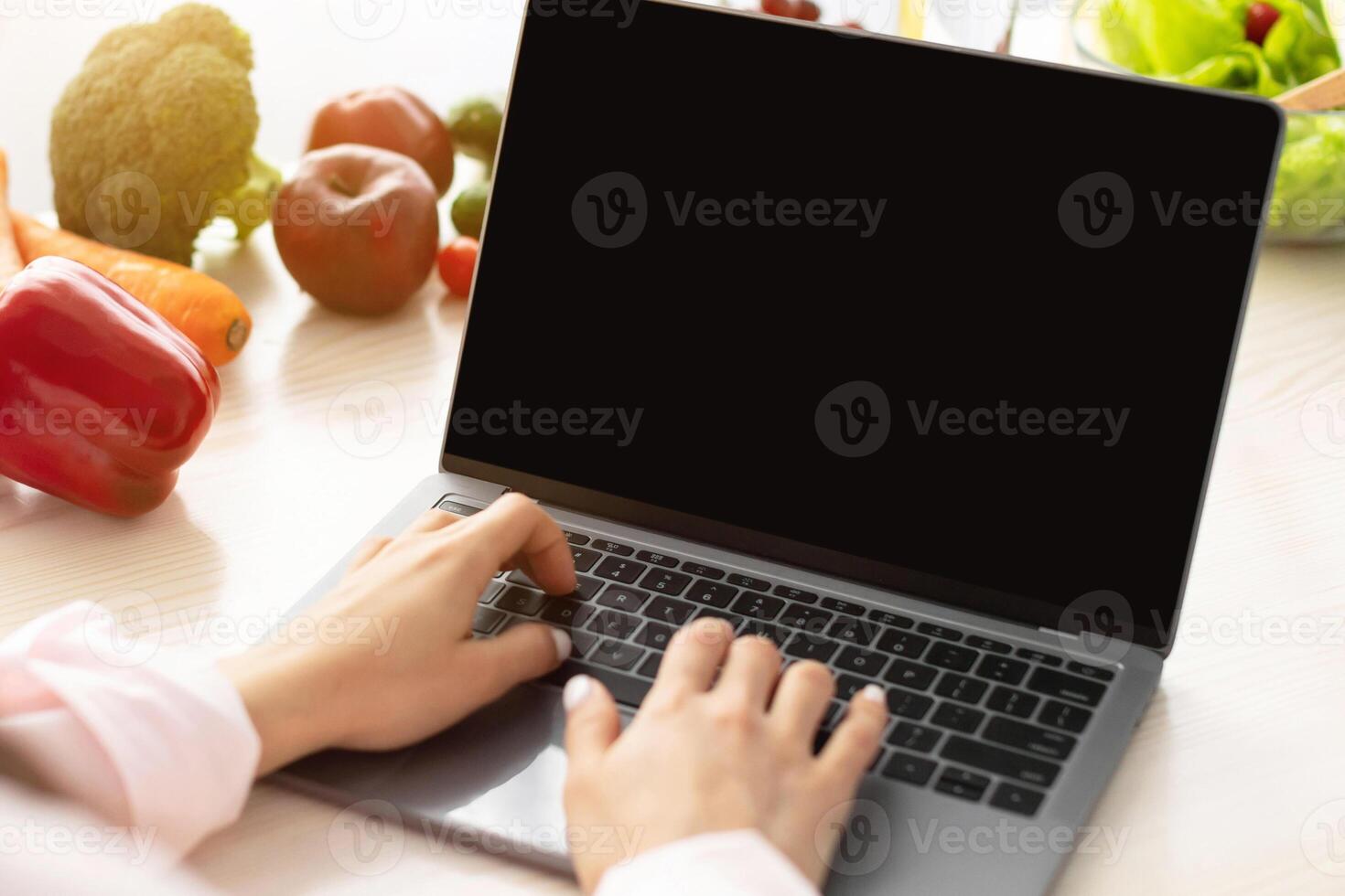 Over the shoulder view of young woman using laptop computer with black empty screen for mockup template, cooking in kitchen, typing on keyboard, searching for vegan recipes online via internet photo