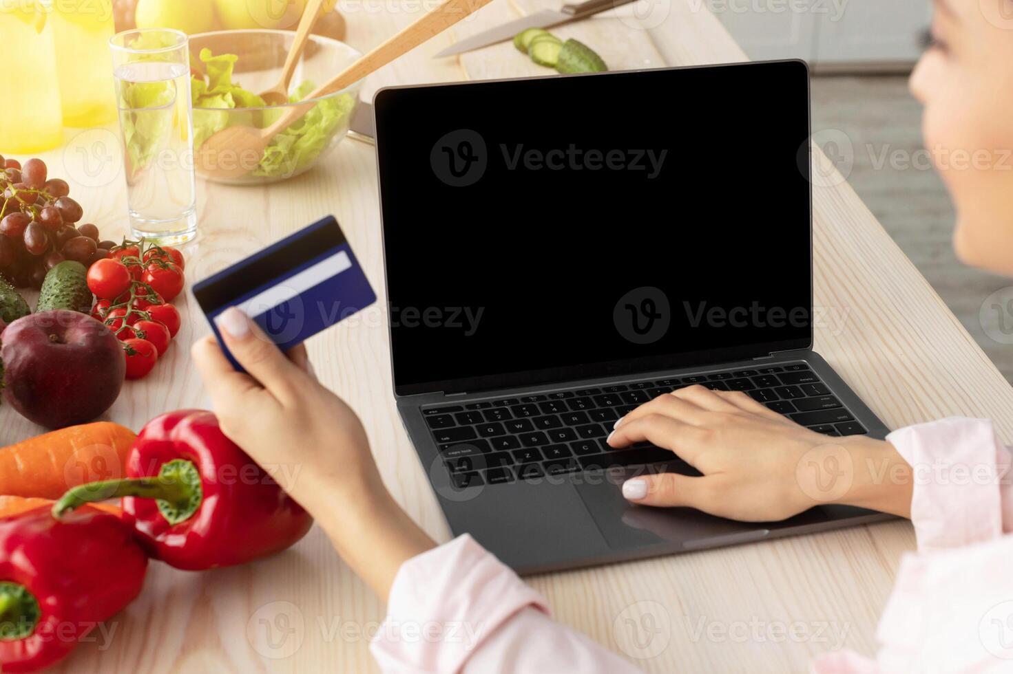 Webshop Concept. Over the shoulder view of unrecognizable positive woman using laptop pc with empty black screen for mock up, sitting at kitchen counter, holding and showing credit card, paying online photo