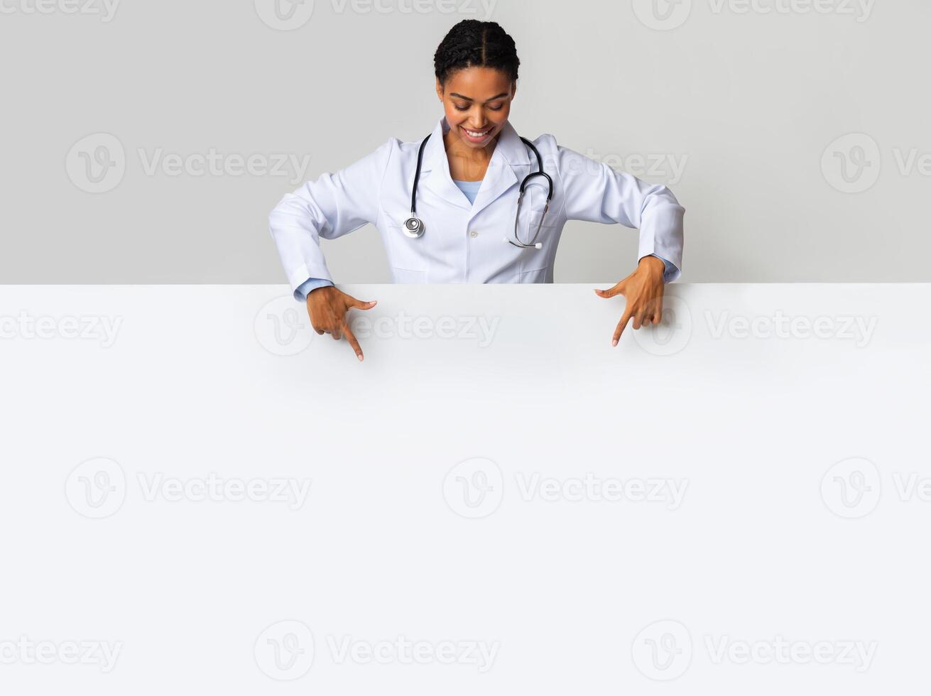 Medical offer. Afro female doctor looking down and pointing at blank white advertising board with empty space for text photo