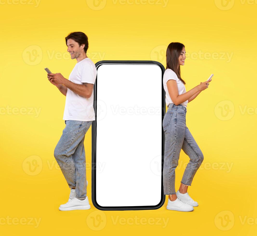 Great App. Excited Couple Leaning On Big Smartphone With Blank White Screen Using Cell Phones, Cheerful Guy And Lady Chatting On Social Media, Standing On Yellow Orange Background, Mock Up, Full Body photo