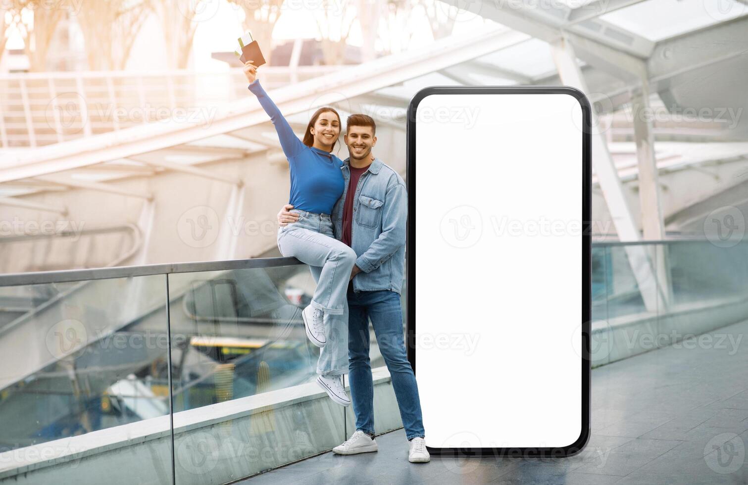 A cheerful couple poses joyfully beside a large blank screen in a modern indoor space. The woman raises her phone, while the man stands beside her with a smile, showcasing a lively moment. photo