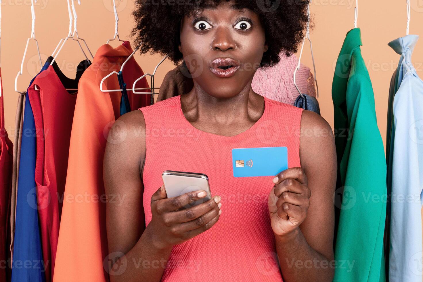 Mobile Shopping Application. Amazed Black Female Holding Cellphone And Credit Card Looking At Camera In Amazement Posing Near Clothing Rack Purchasing Clothes Online On Yellow Background In Studio photo