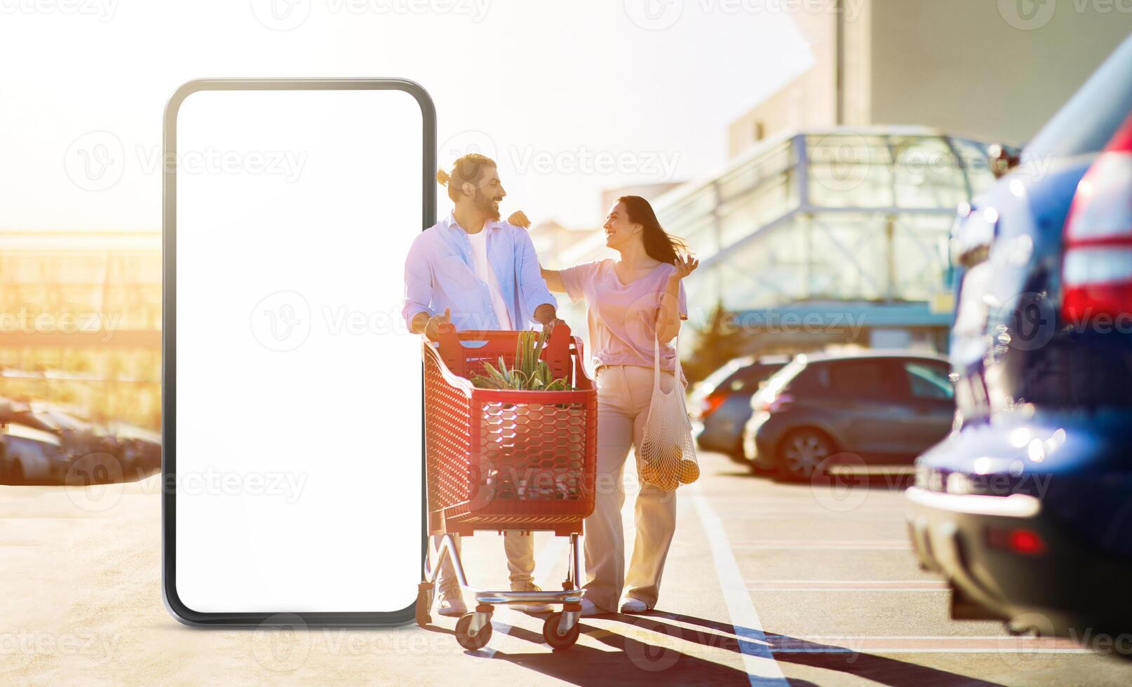A couple enjoys their time shopping together, smiling and interacting as they push a cart filled with groceries. The sun casts a warm glow on the bustling parking lot with huge phone with blank screen photo