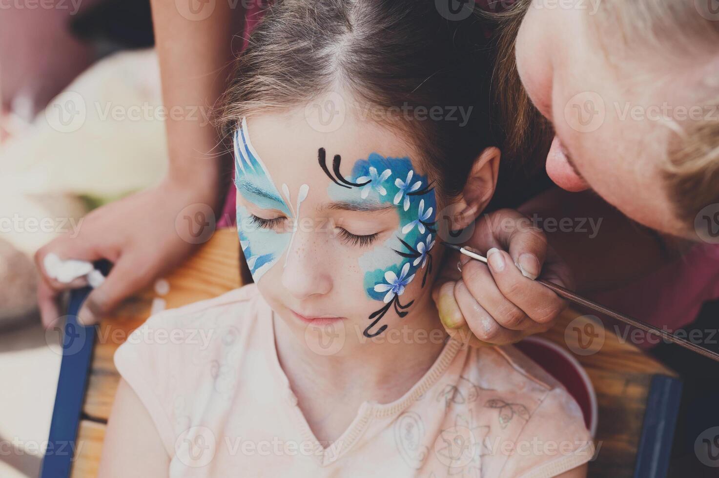 Child animator, artist's hand draws face painting to little girl. Child with funny face painting. Painter makes blue butterfly at girl's face. Children holiday, event, birthday party, entertainment. photo