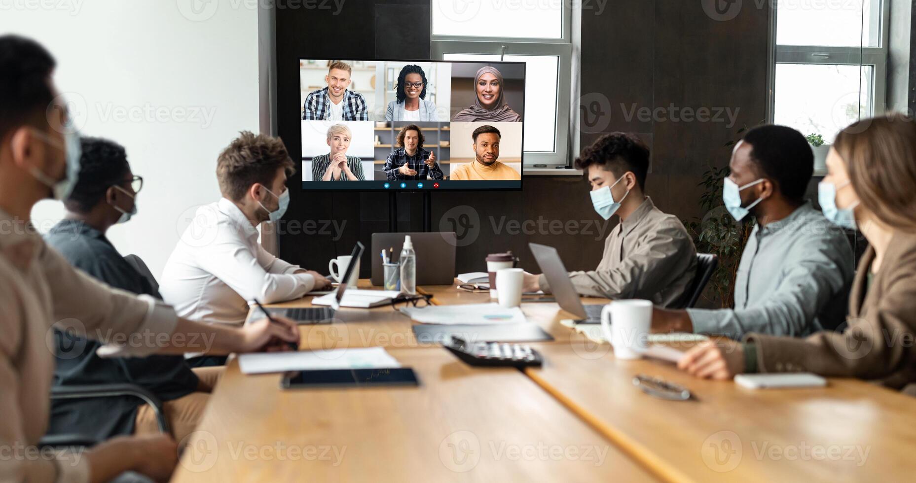 Videoconferencing, Virtual Meeting. Diverse group of workers in masks making online videocall with colleagues, sitting at table in boardroom, looking at screen, talking on web with remote employees photo