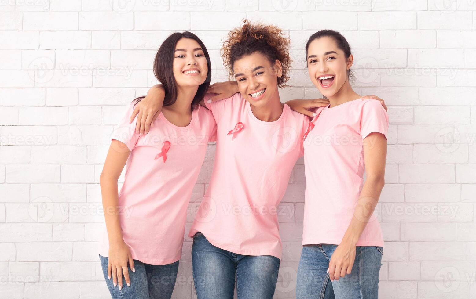 Happy Diverse Ladies In Pink Breast Cancer T-Shirts Hugging Posing Over White Brick Wall Background. Oncology Awareness Concept photo