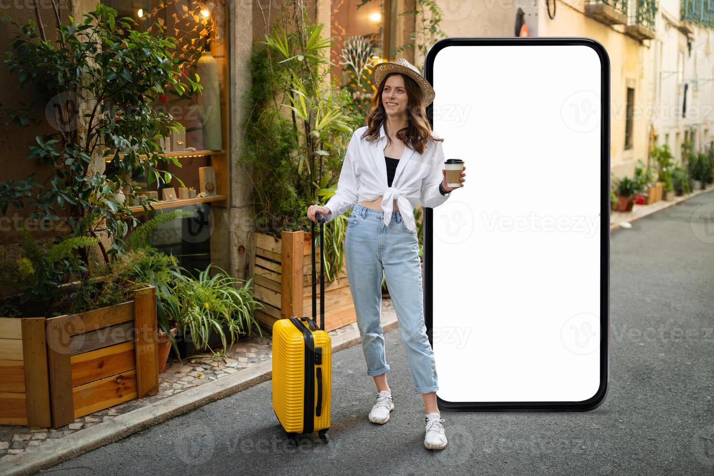 A traveler dressed in a stylish outfit holds a coffee cup while standing beside a bright yellow suitcase. The person poses in a charming street surrounded by greenery and buildings. photo
