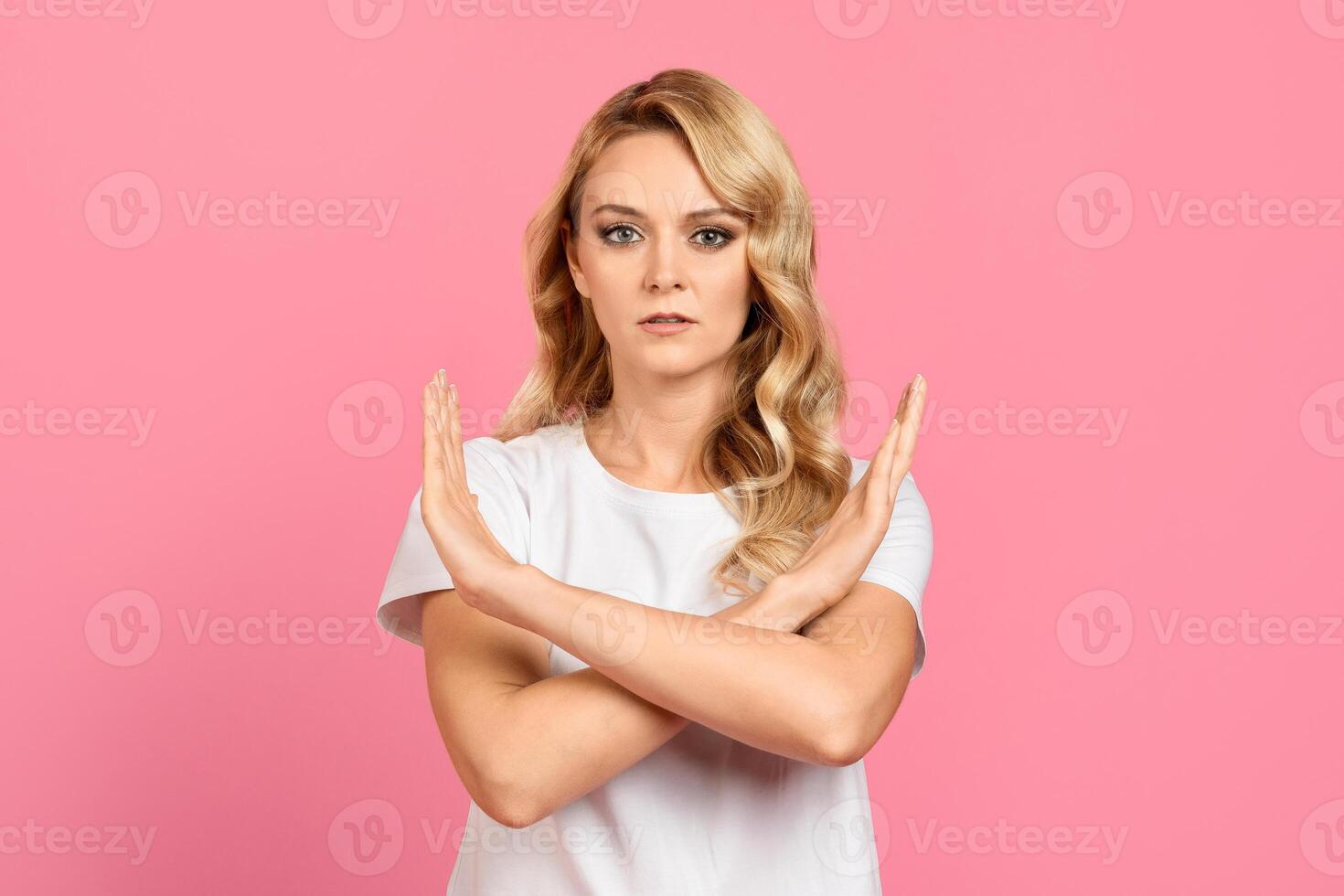 A woman with wavy blonde hair stands confidently, arms crossed in a stop gesture. She is wearing a white top and has a serious expression on her face, set against a vibrant pink backdrop. photo