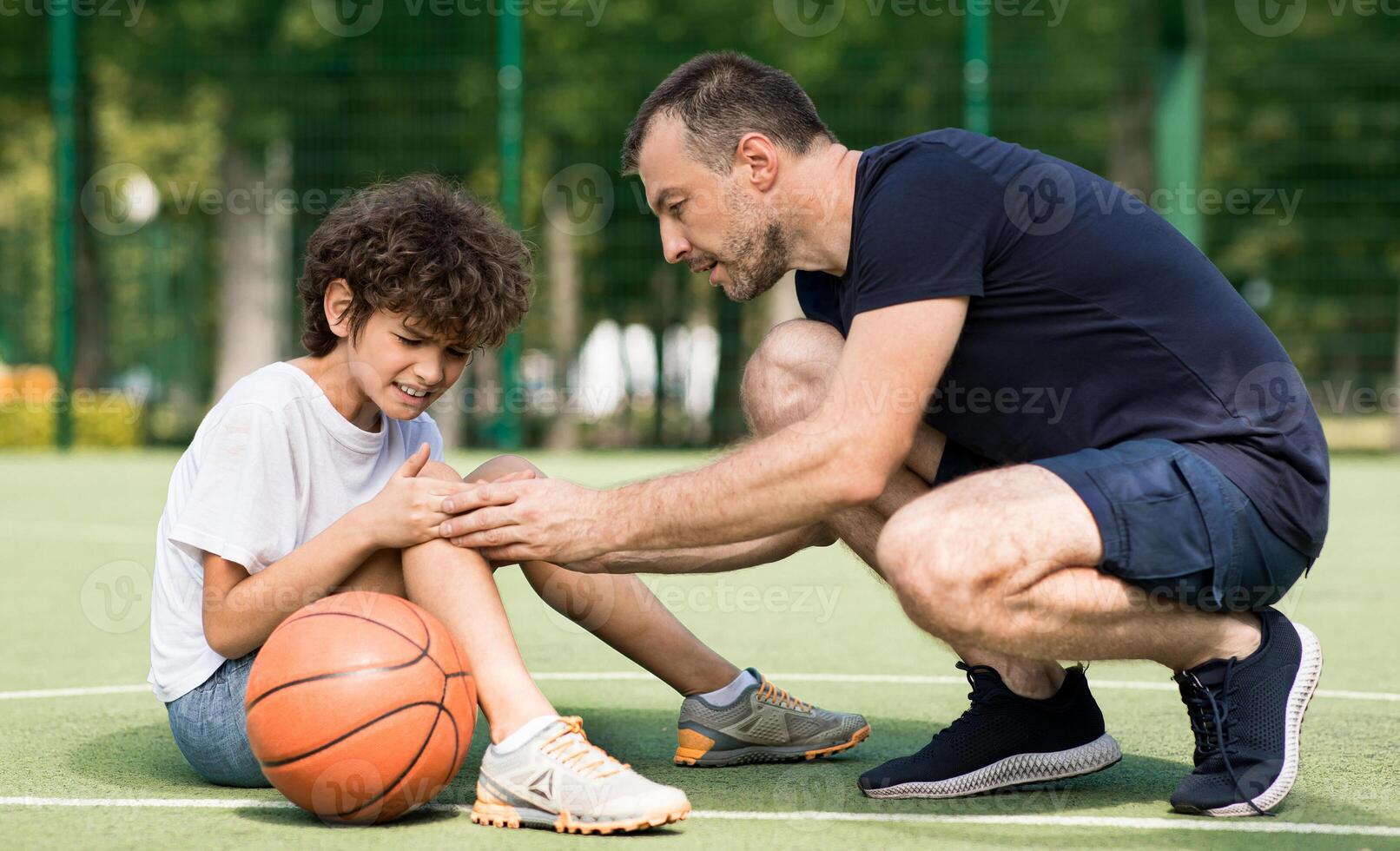 Side view portrait of focused adult man coach helping boy with knee trauma after playing basketball on the court, sad player feeling pain, touching leg. Activity And Sports Injury Concept. photo